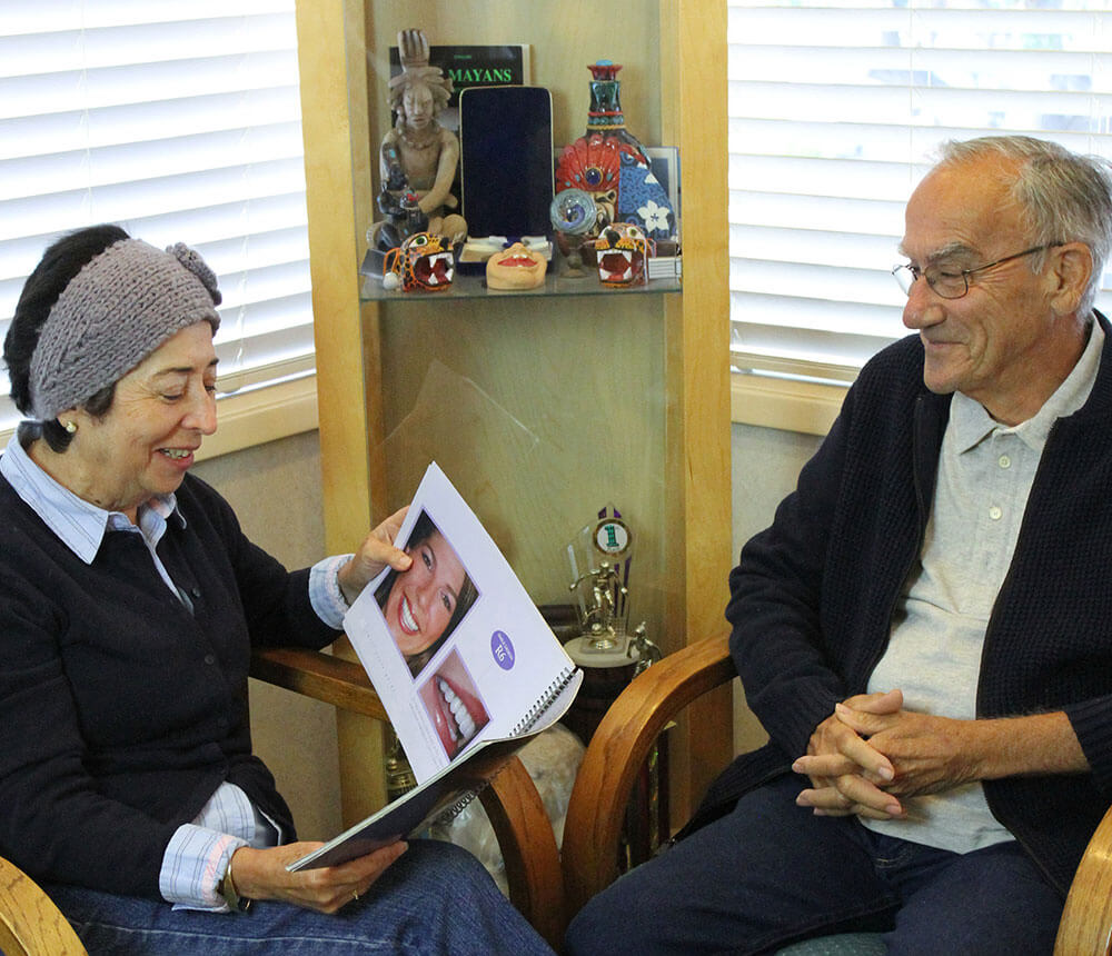 A woman and a man sit in chairs, angled toward each other, while the woman looks a book of smiling patients