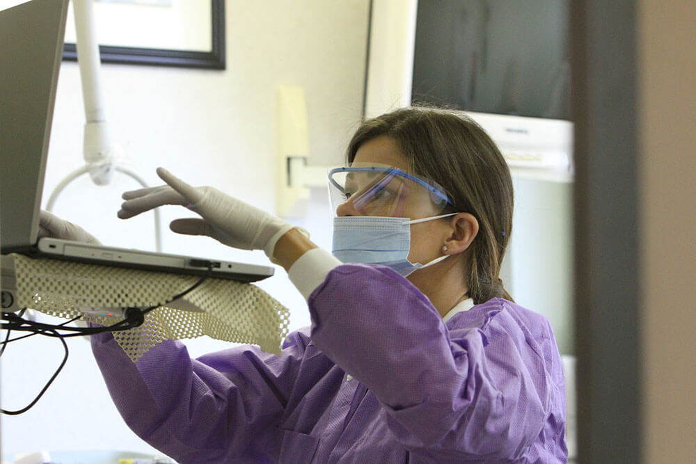 A dental assistant, in a purple gown, googles and face mask, typing on a laptop with gloved hands
